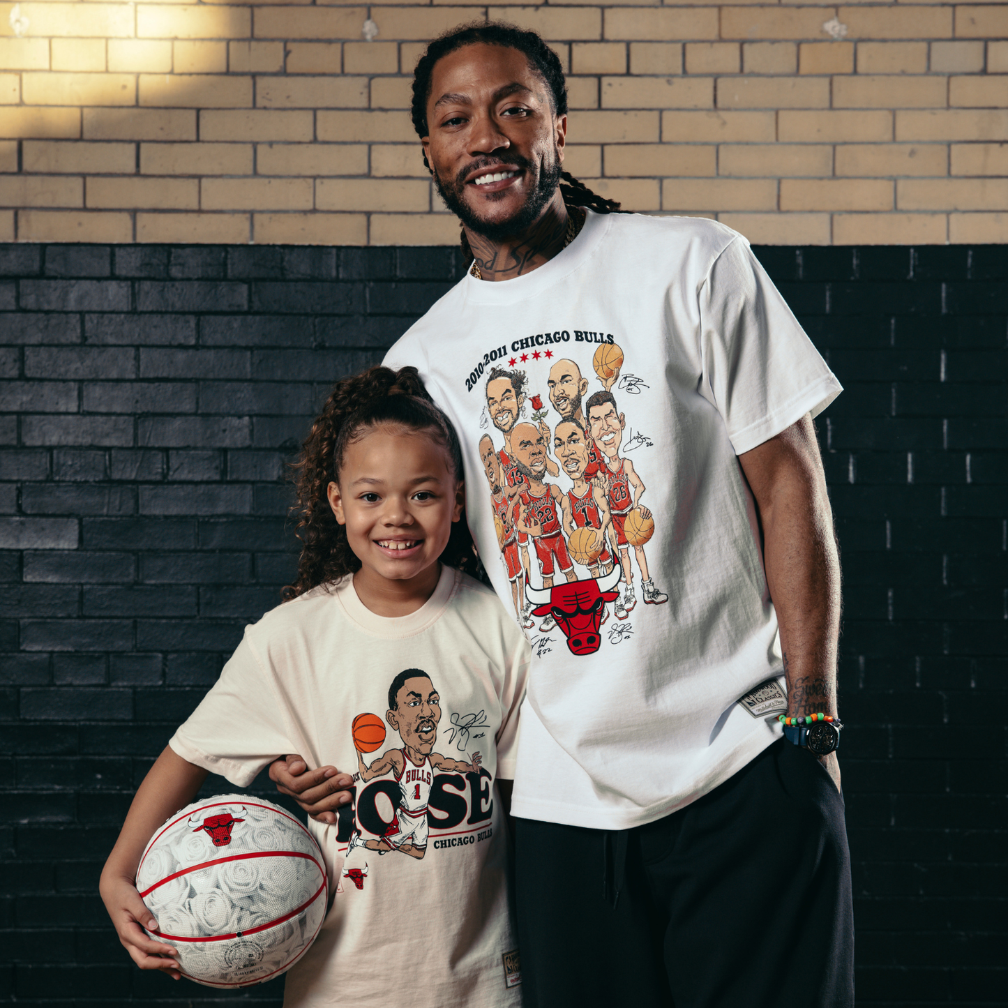 Young girl wearing t-shirts with sports-themed graphics, holding a basketball, against a brick wall.